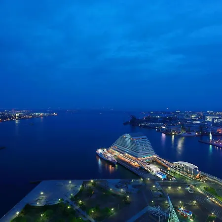 Hotel Okura Kobe Exterior photo The photo shows a serene evening view of a waterfront cityscape. The scene is illuminated by soft artificial lighting, creating a tranquil ambiance. In the foreground, there are landscaped areas with greenery, and a waterfront promenade lined with li