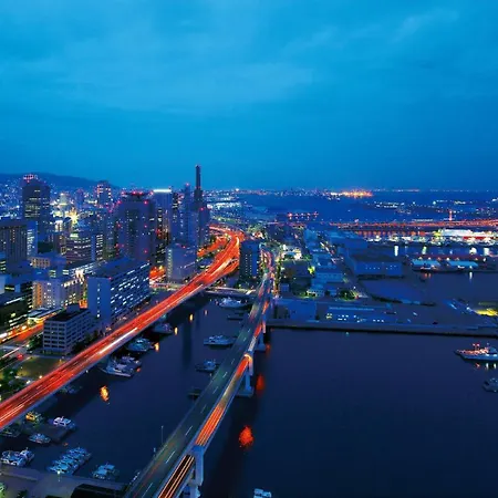 Hotel Okura Kobe Exterior photo The photo depicts a cityscape at dusk, showcasing a vibrant urban environment. The skyline features tall buildings illuminated by city lights, creating a glowing effect against the twilight sky. A river or harbor is visible, with boats docked along t